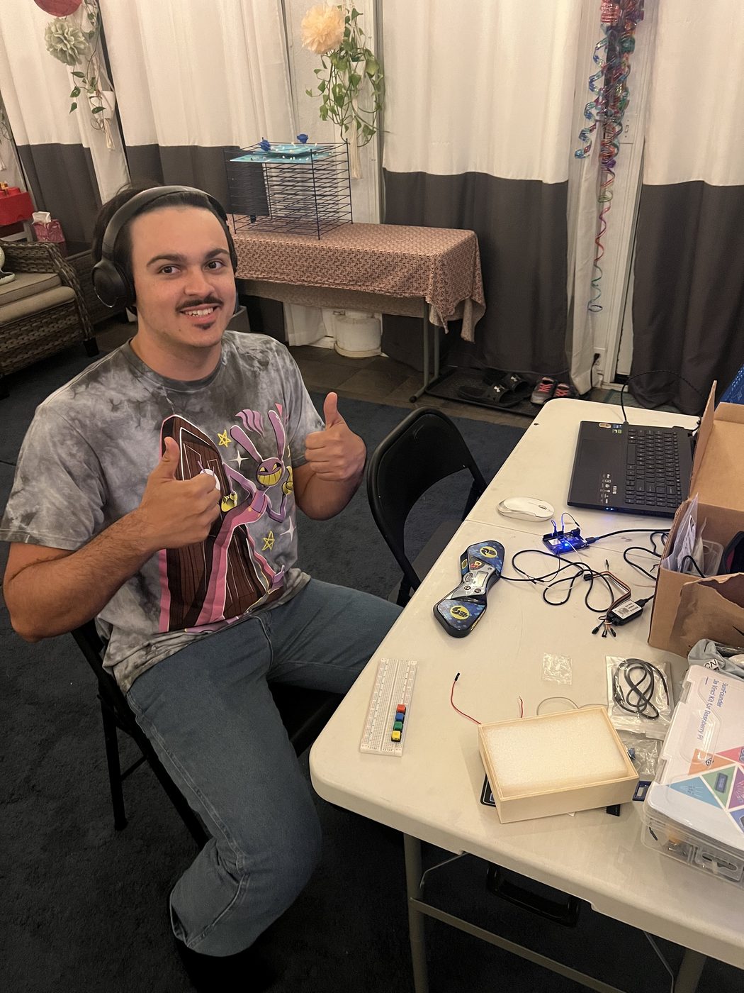 Roger Jr. at the workbench giving a thumbs up, with a laptop, a StackMat timer and prototype electronics on the table.